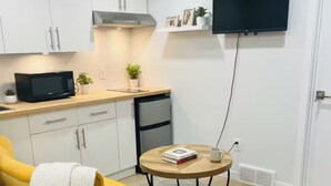 Una cocina compacta con armarios blancos, un microondas y un refrigerador pequeño. Hay un sill ón amarillo con una manta blanca, una mesa redonda de madera con un libro y una taza, y un televisor de pantalla plana montado en la pared.