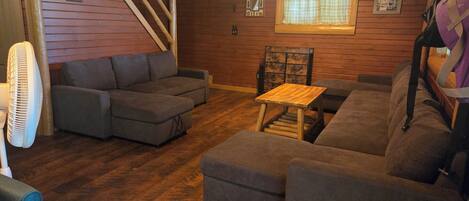 A cozy living room with a wooden floor, grey sofas, a wooden coffee table, and a fan.
