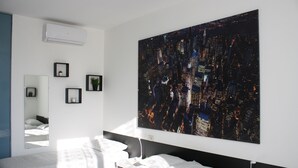 Una camera d'albergo moderna con un'ampia pittura raffigurante un panorama urbano, un letto con una coperta a motivi decorativi, un comodino con una lampada e una piccola pianta in vaso.