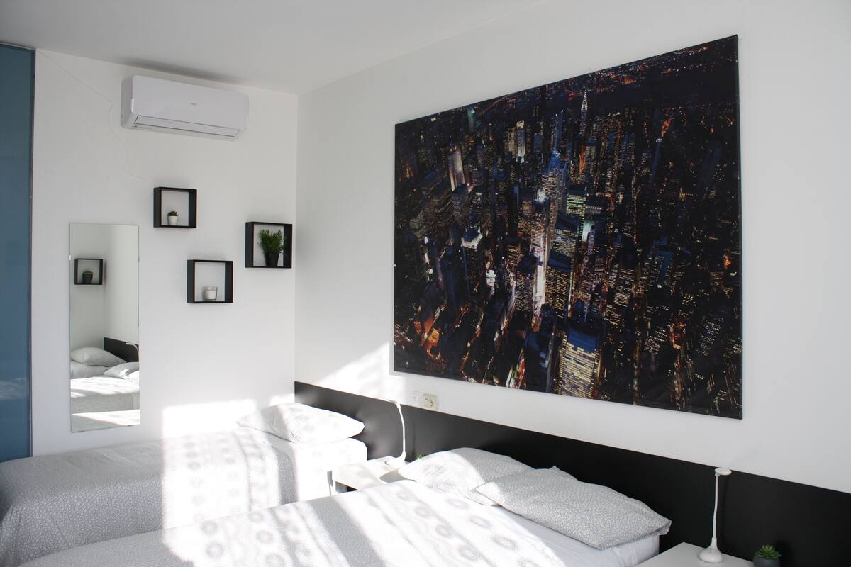 A modern hotel room with a large cityscape painting on the wall, a bed with a patterned bedspread, a bedside table with a lamp, and a small potted plant.