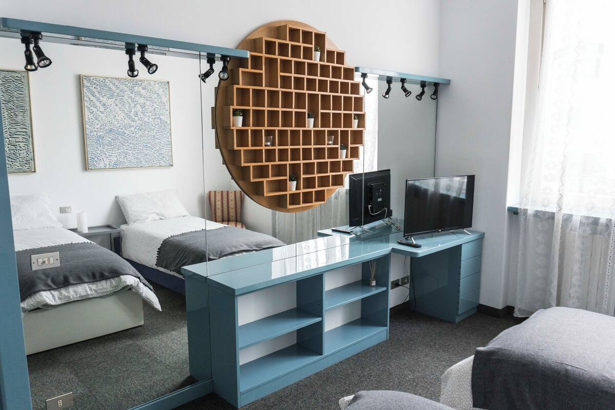 A bedroom with a bed, a TV, a desk, a mirror, and a wall-mounted shelf.