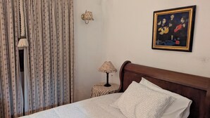 Un dormitorio con una cama de madera, cortinas con estampado floral, una mesita de noche con lámpara y un cuadro en la pared.