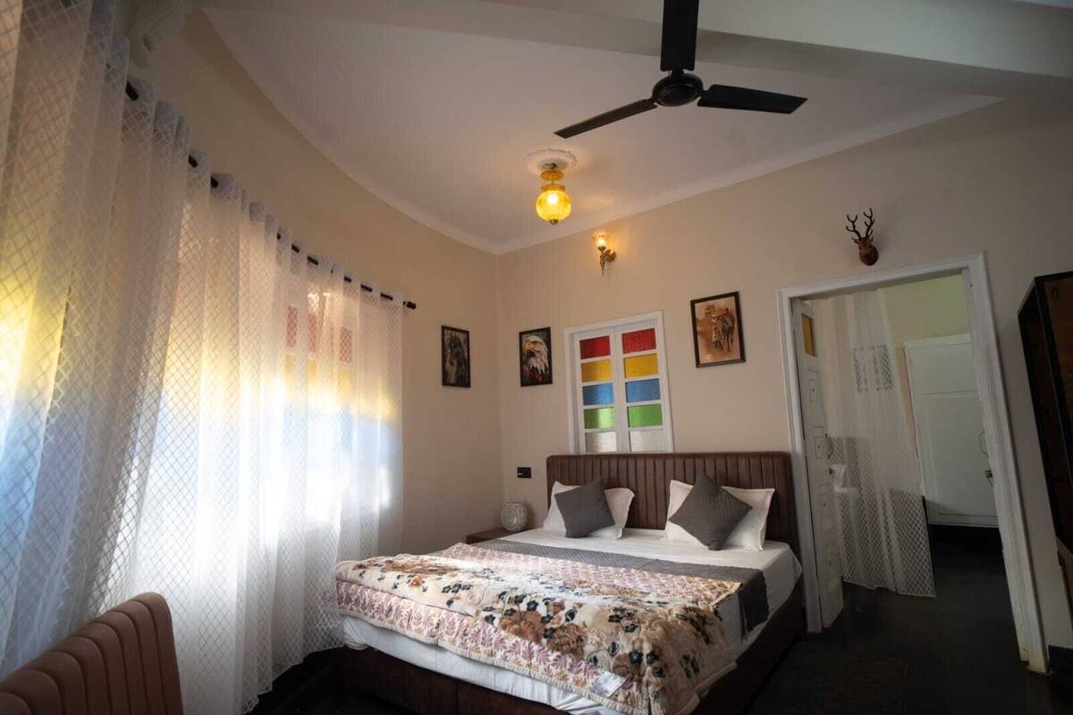 Une chambre à coucher comprenant un lit, un ventilateur de plafond, une lampe fixée au mur, une porte et une fenêtre avec des rideaux légers.