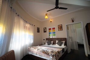 A bedroom with a bed, a ceiling fan, a wall-mounted light, a door, and a window with sheer curtains. - L2 Darbar Kothi (Jodhpur)