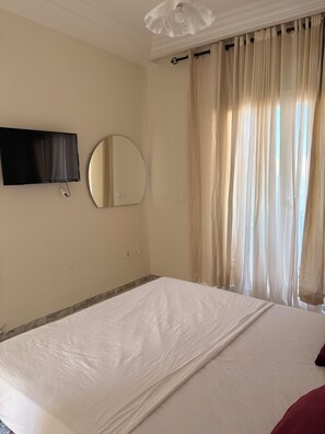 A hotel room with a bed, a television, a round mirror, and curtains. - A cozy little apartment just a stone's throw from the ocean (Hammamet)