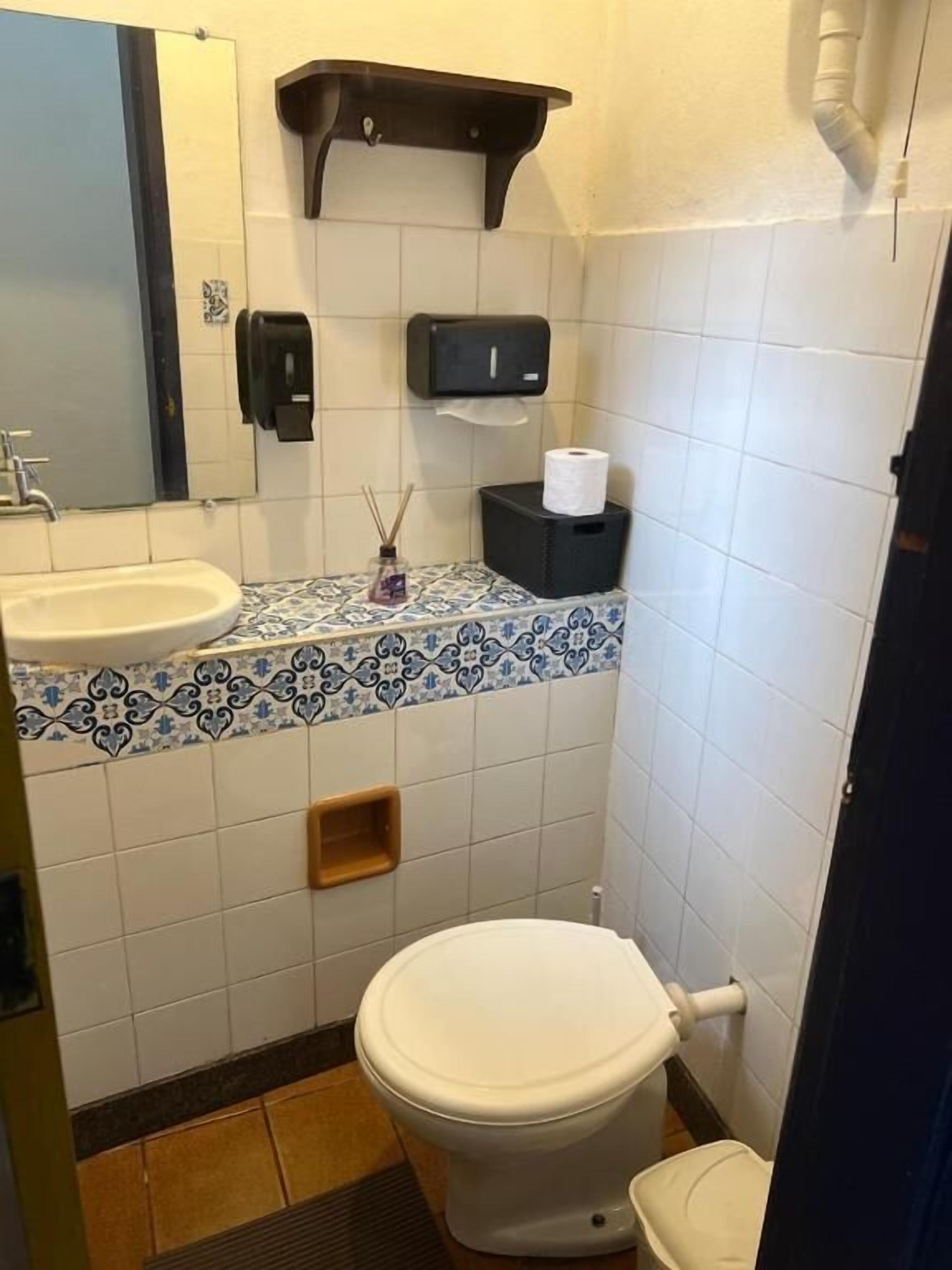 A bathroom with a toilet, sink, mirror, towel dispenser, and a small shelf.