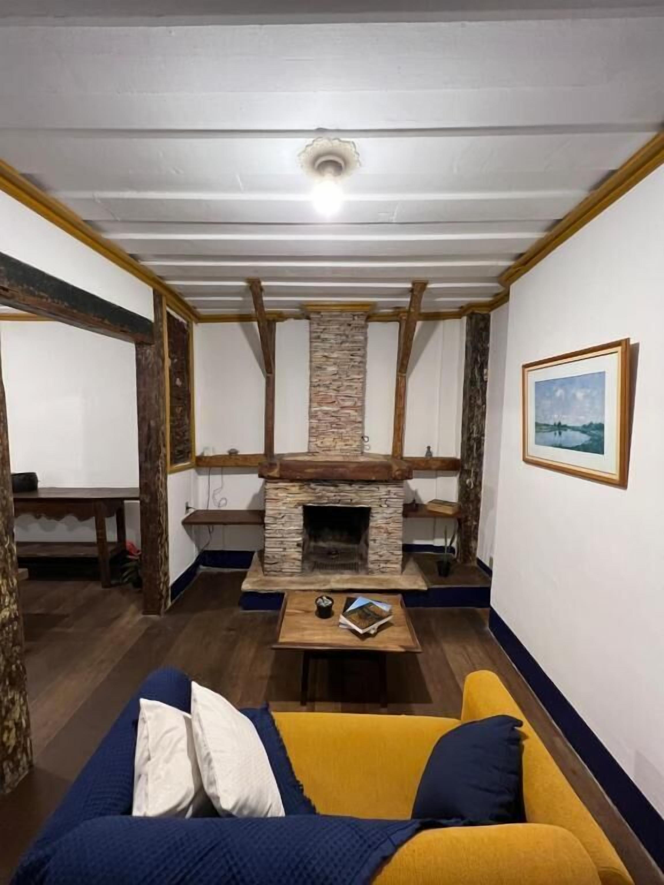 Ett rum med en steneldstad, träbjälkar, en gul soffa och en inramad tavla på väggen.