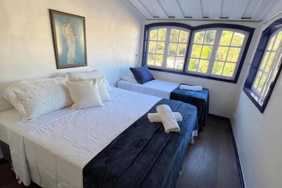 A room with a large bed, a smaller bed, and a bench. There are windows with views of greenery.