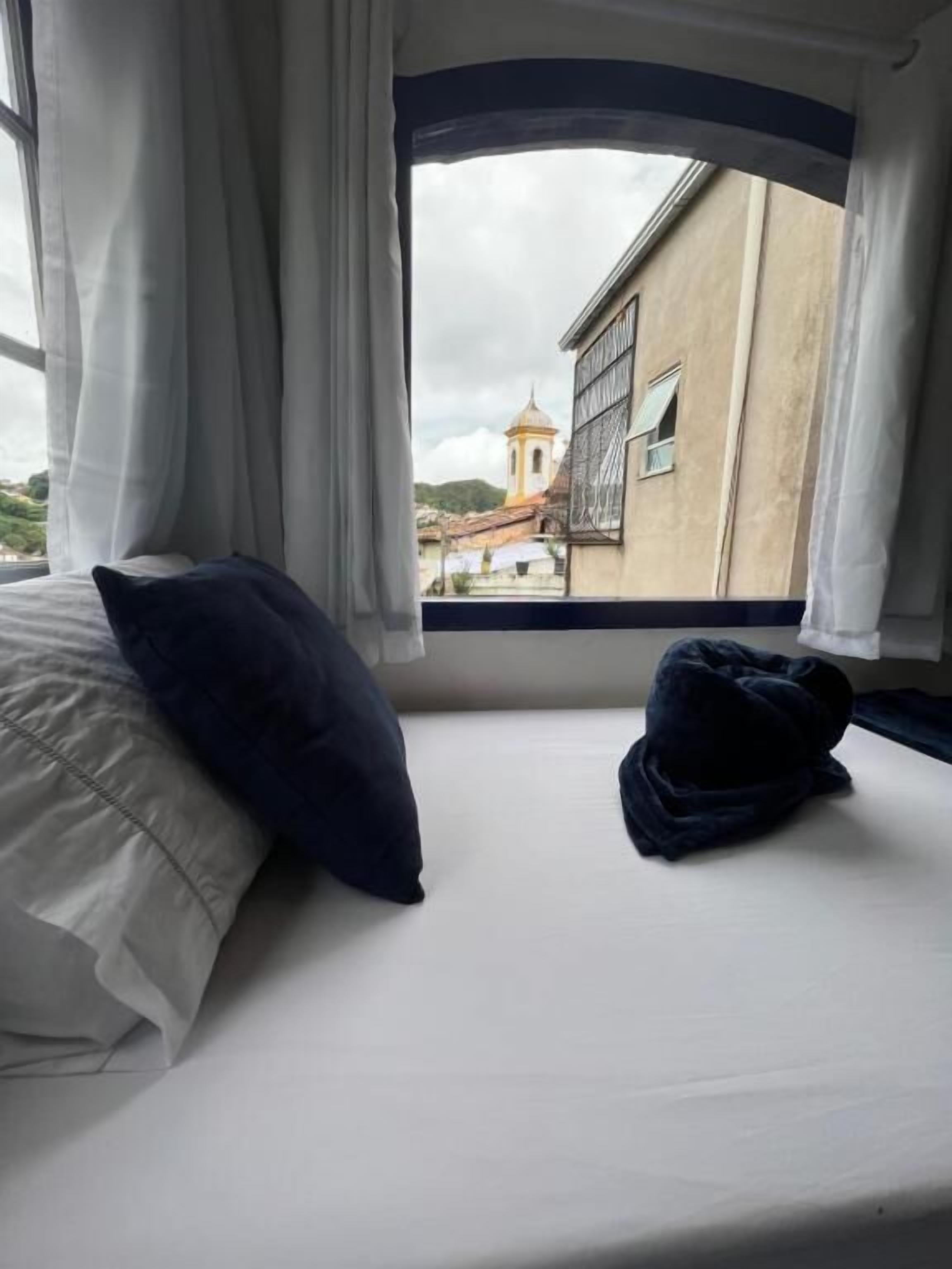A bed with white and dark blue pillows, a window with white curtains, and a view of a building with a bell tower.