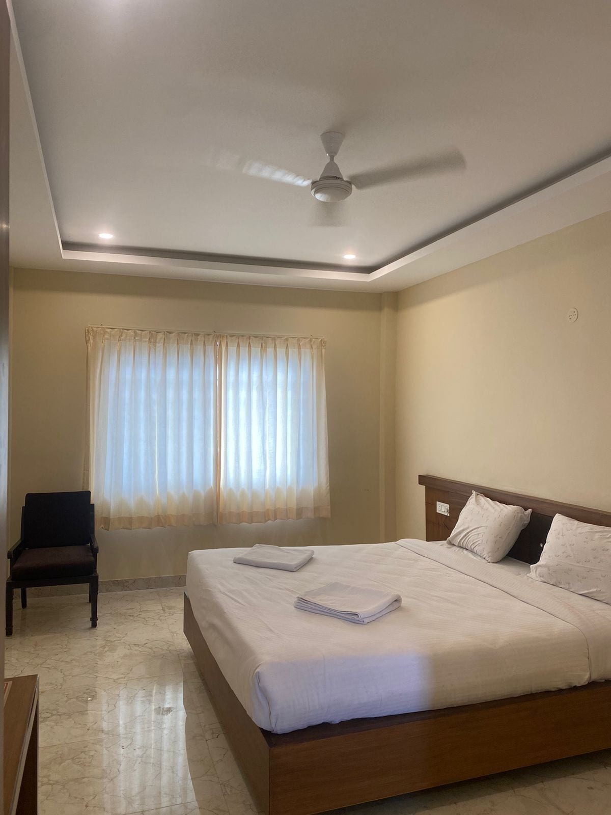 A hotel room with a large bed, a chair, a ceiling fan, and a window with curtains.