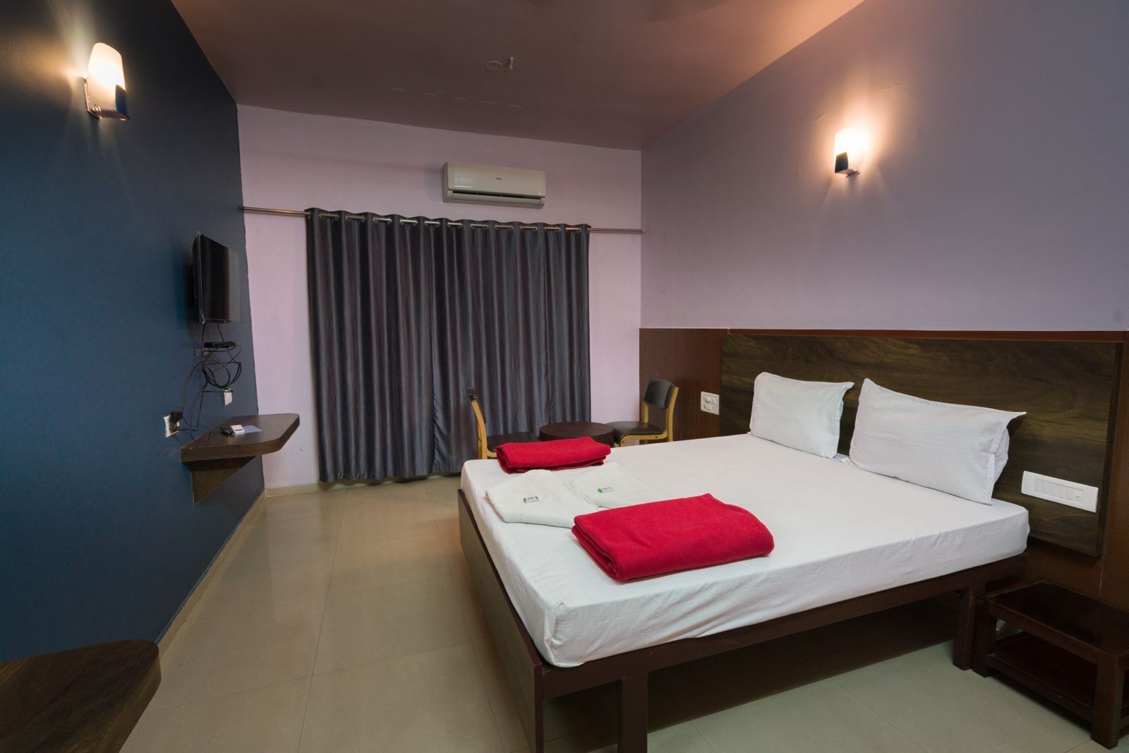 A hotel room with a bed, a desk, a chair, a TV, and an air conditioning unit.