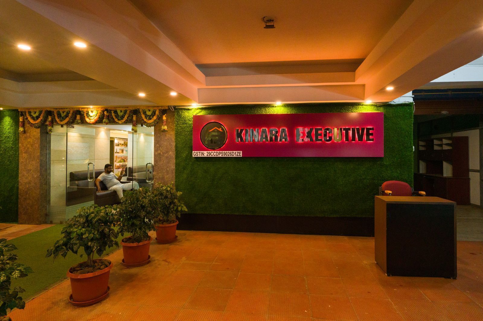 A reception area with a sign reading "KINARA EXECUTIVE" and a GSTIN number. There are potted plants and a reception desk.