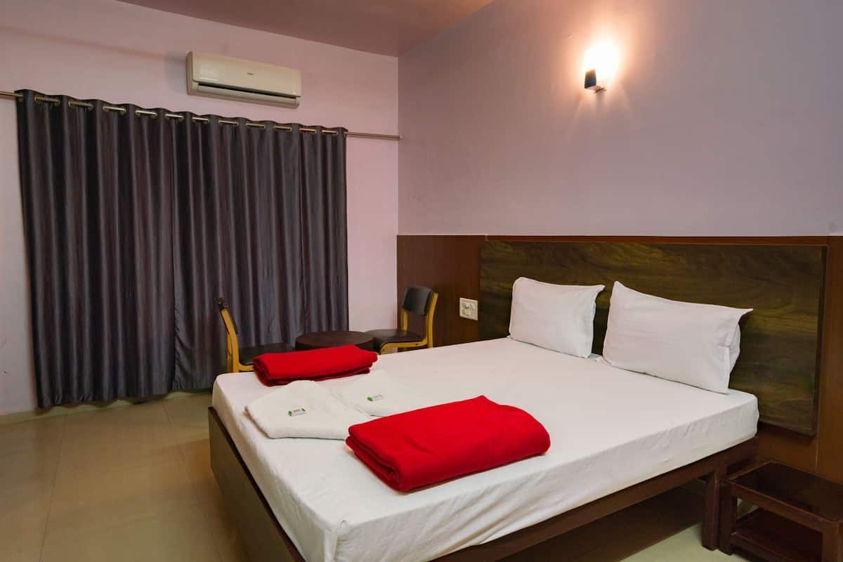 A hotel room with a bed, a chair, a small table, a wall-mounted air conditioner, and a wall-mounted light fixture.