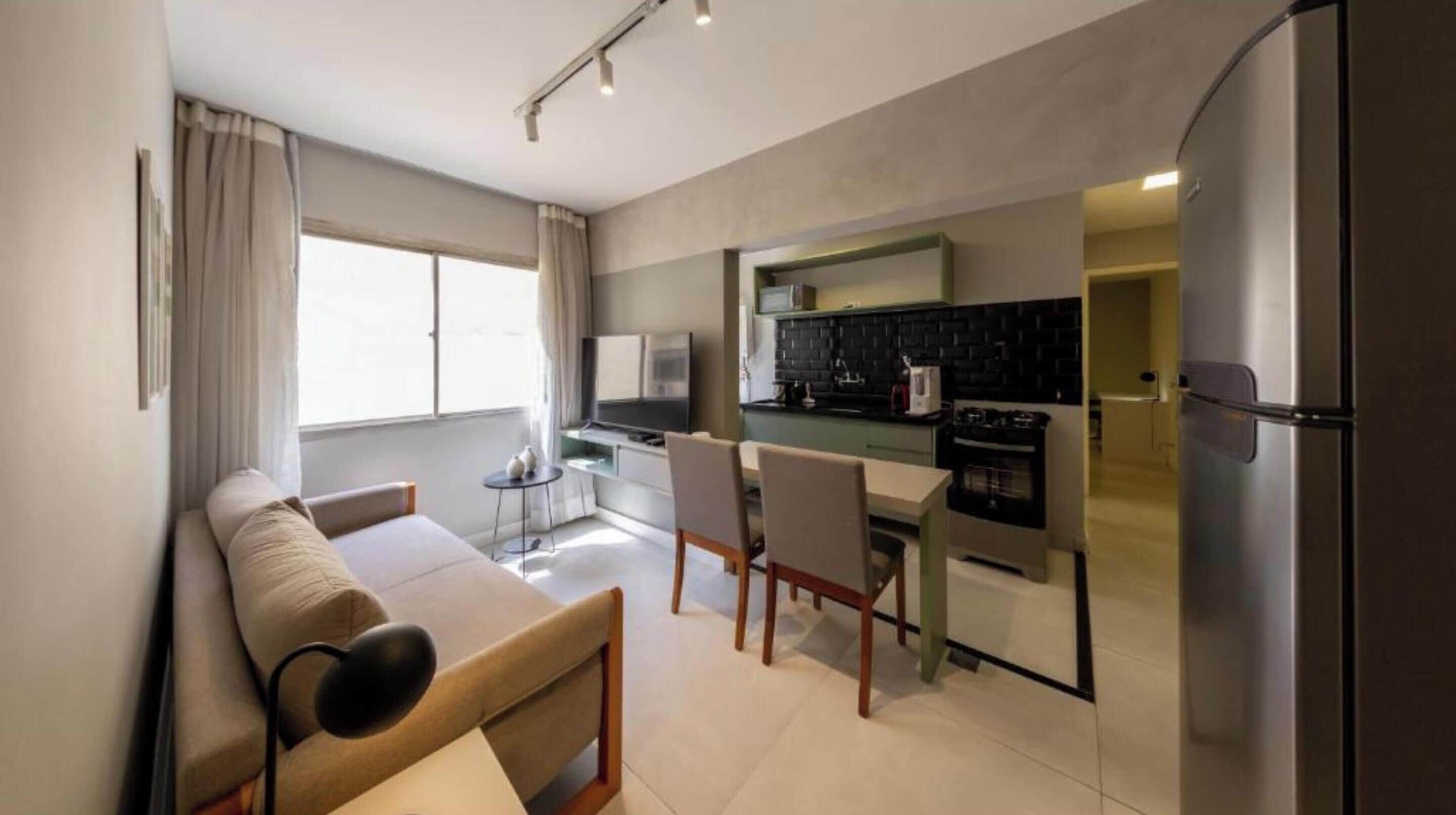 A modern living room with a sofa, a small table, and a TV. The kitchen area includes a stove, a refrigerator, and a dining table with chairs.