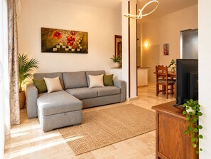A living room with a grey sectional sofa, a woven rug, a flat-screen TV, and a painting on the wall. - Capo Comino 1 - Two Bedroom Apartment, Sleeps 5 (La Caletta)