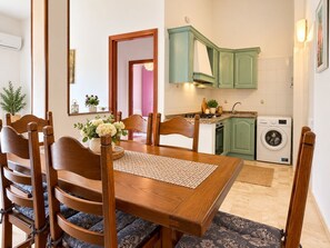 Een eethoek met een houten tafel en stoelen, een keuken met groene kastjes, en een wasmachine.