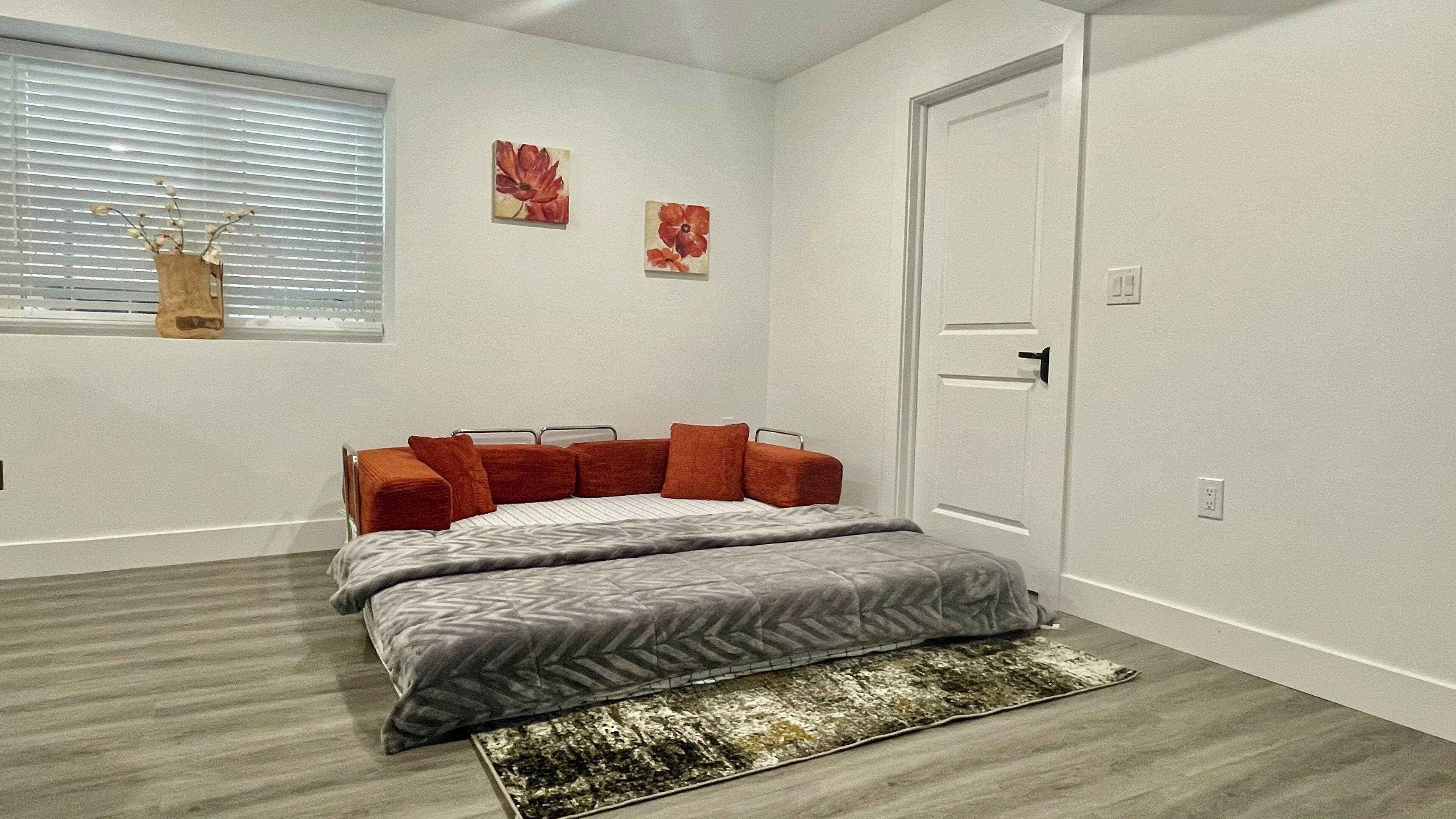 A bedroom with a bed, a window with blinds, a door, and two wall-mounted flower paintings.