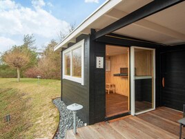A modern building with a dark exterior, a wooden deck, and a glass door leading to a room with a wooden interior.