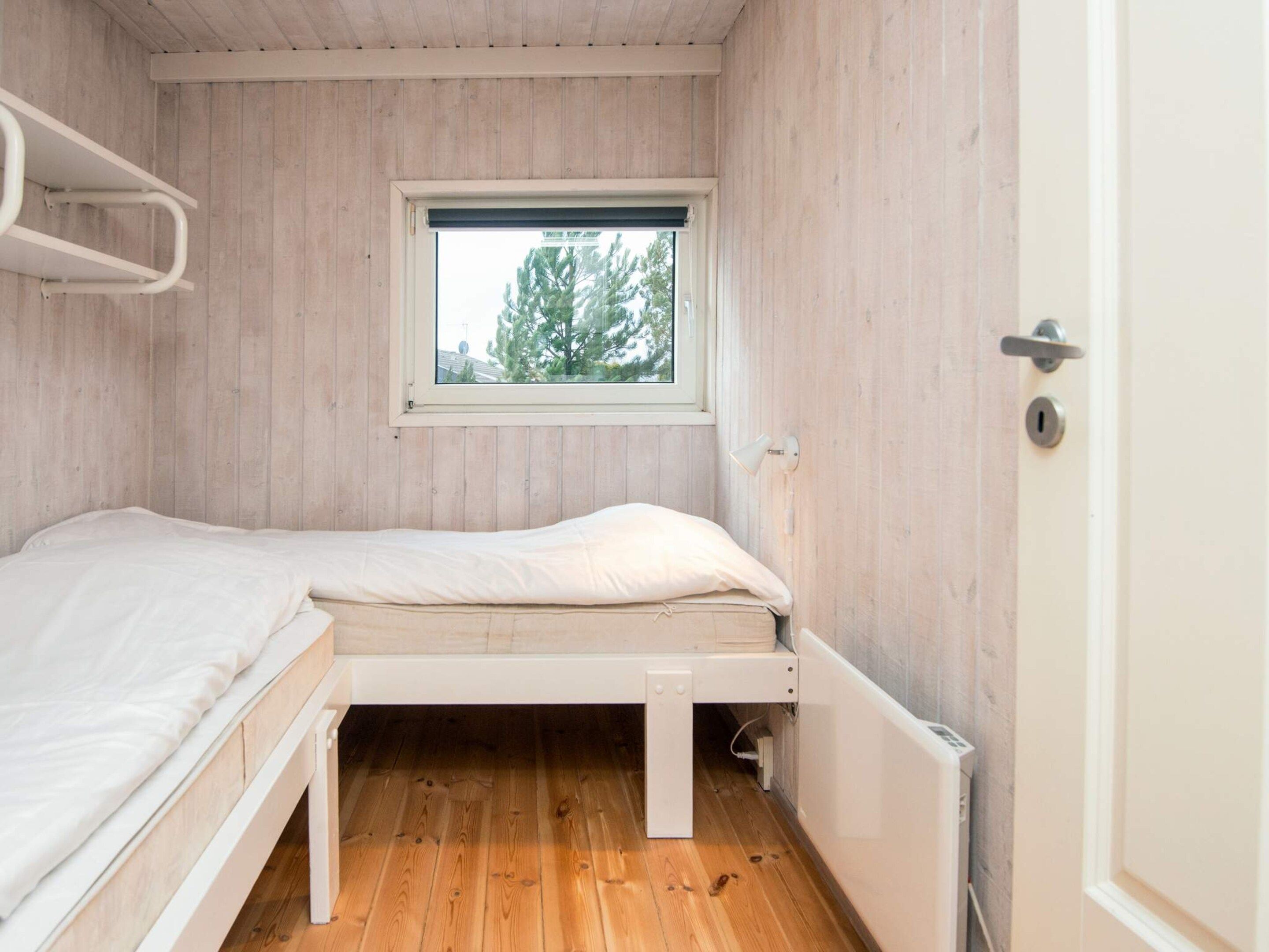 A small, single-bed room with a wooden floor, a window with a view of trees, and a white door.