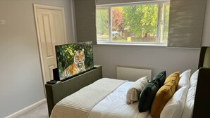 A bedroom with a bed, a television on a stand, a window with a view of trees, and a door. - Home in Milton Keynes (Milton Keynes)