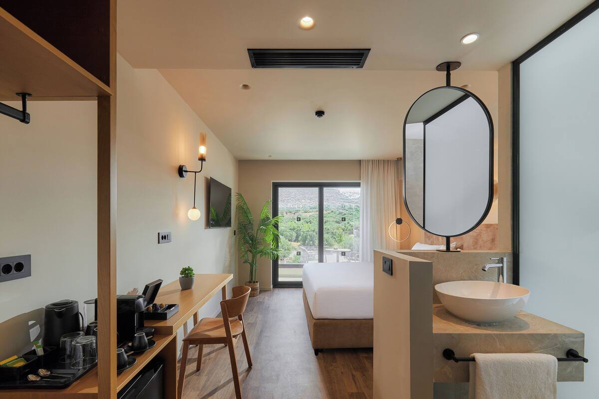 Una habitación de hotel moderna con una cama, un escritorio con una silla, un ventanal grande, un baño con lavamanos y espejo, y una planta en maceta.