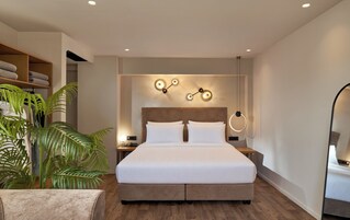 Quarto de hotel moderno com uma cama grande, cabeceira de madeira, luminária fixada na parede e uma planta em vaso.