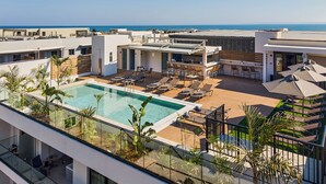 Una piscina en la azotea con sillones de descanso y un área de bar, rodeada de vegetación y con vistas al mar.