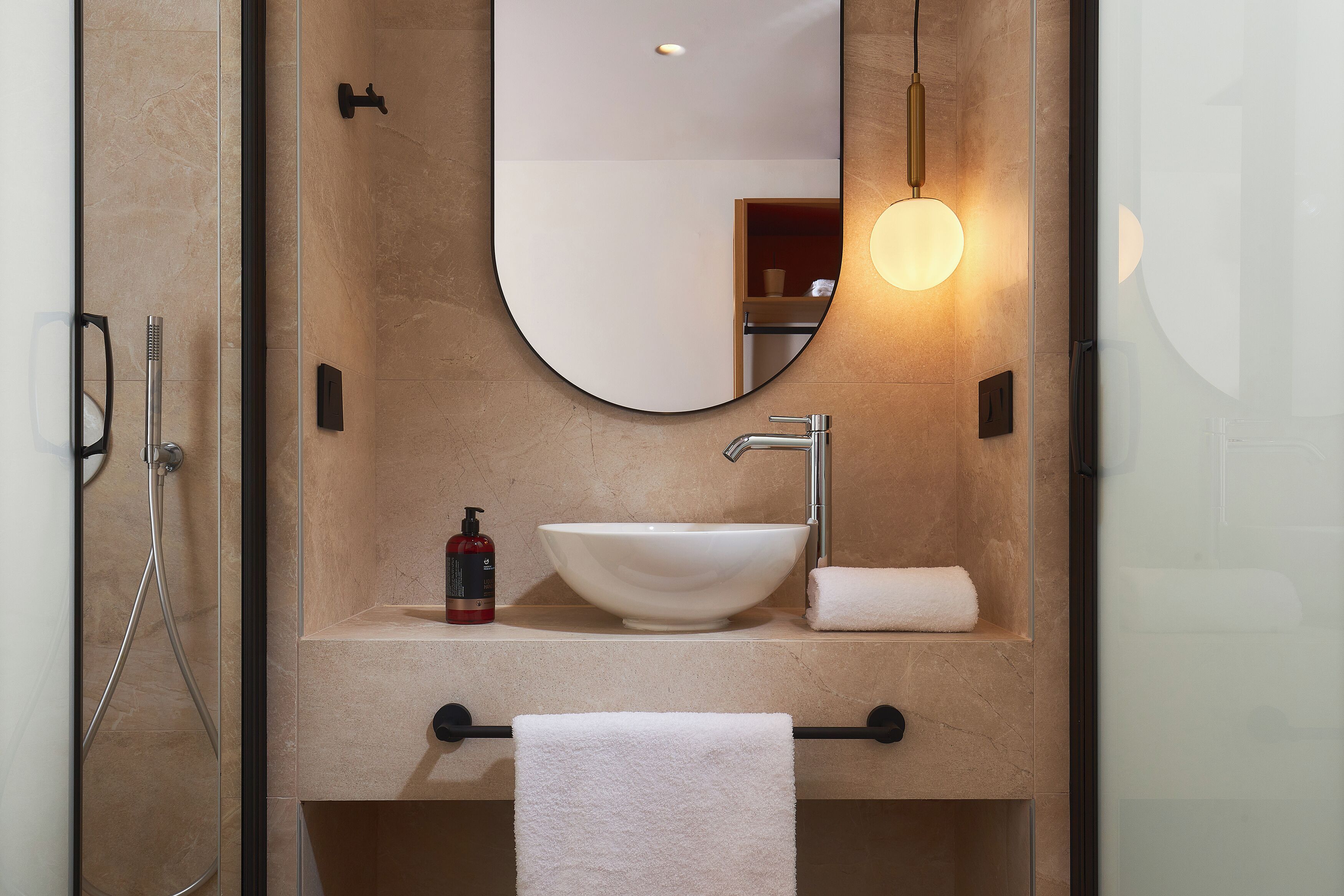 A bathroom with a large oval mirror, a white sink, a towel rack, and a handheld showerhead.