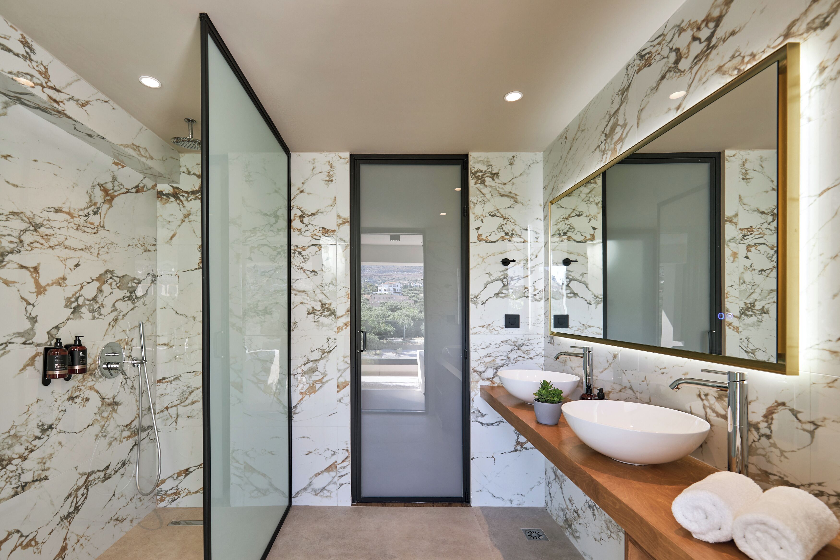 Une salle de bain moderne dotée d’une douche à parois de verre, d’un mur recouvert de carreaux de marbre, d’un grand miroir et d’un meuble-lavabo en bois avec un vasque blanche.