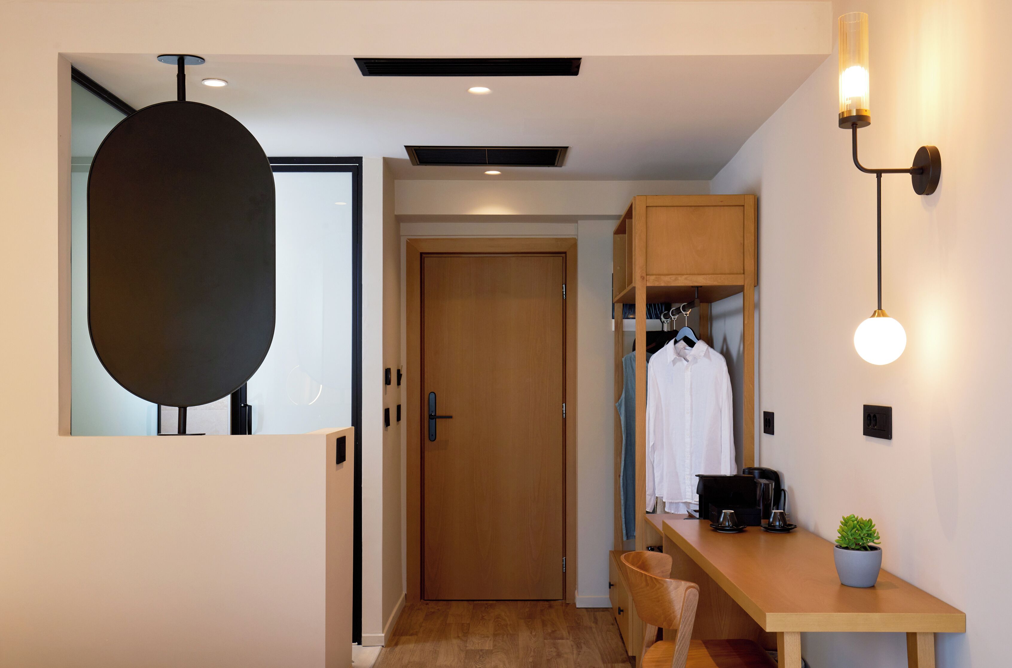 A modern hotel room with a wooden door, a desk with a coffee maker, a hanging light fixture, and a potted plant.