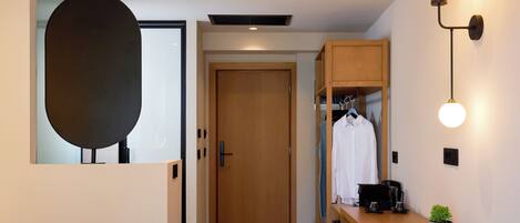 A modern hotel room with a wooden door, a desk with a coffee maker, a hanging light fixture, and a potted plant.