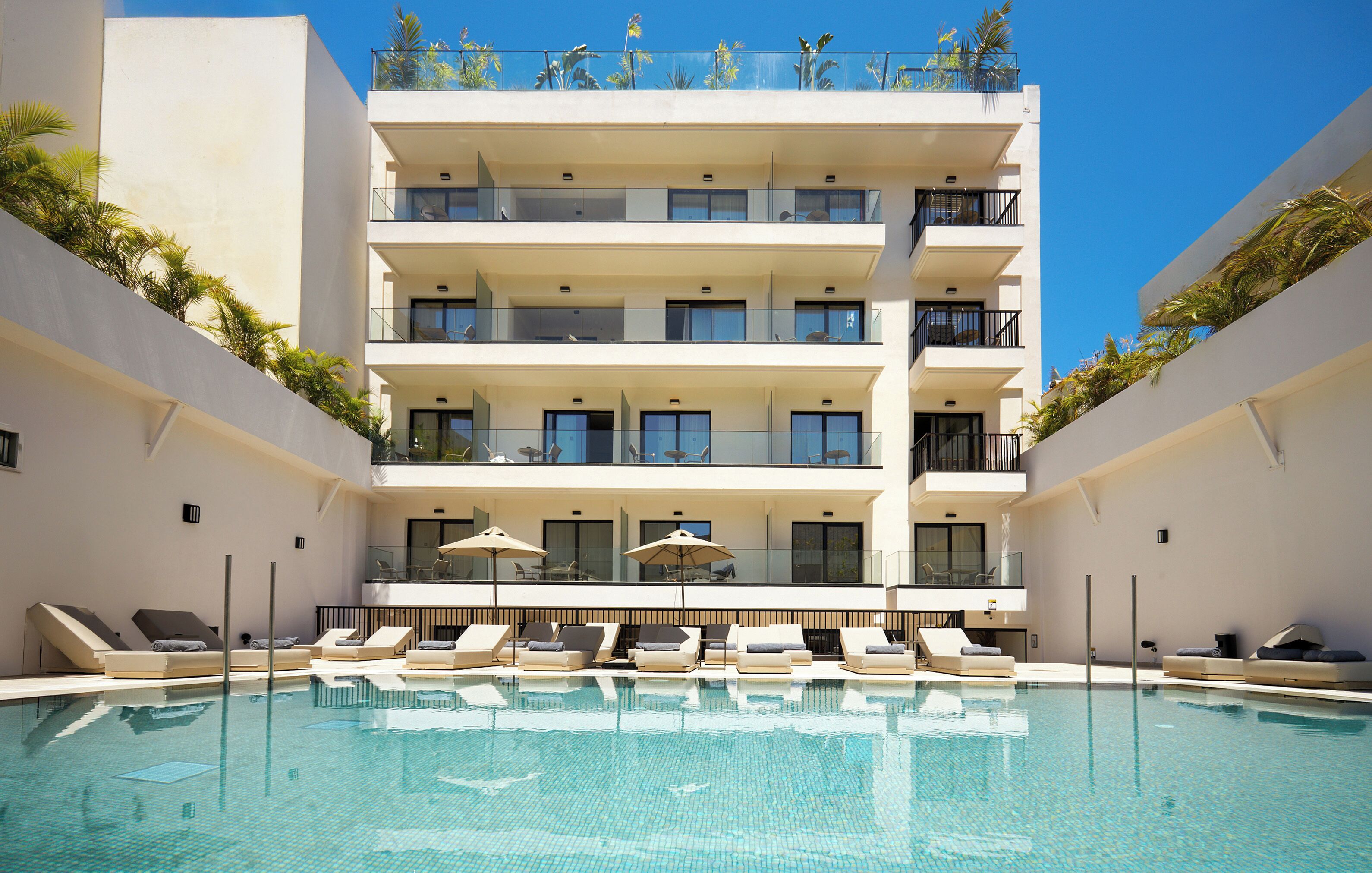 Un hotel de varios pisos con piscina, tumbonas y un balcón con plantas.