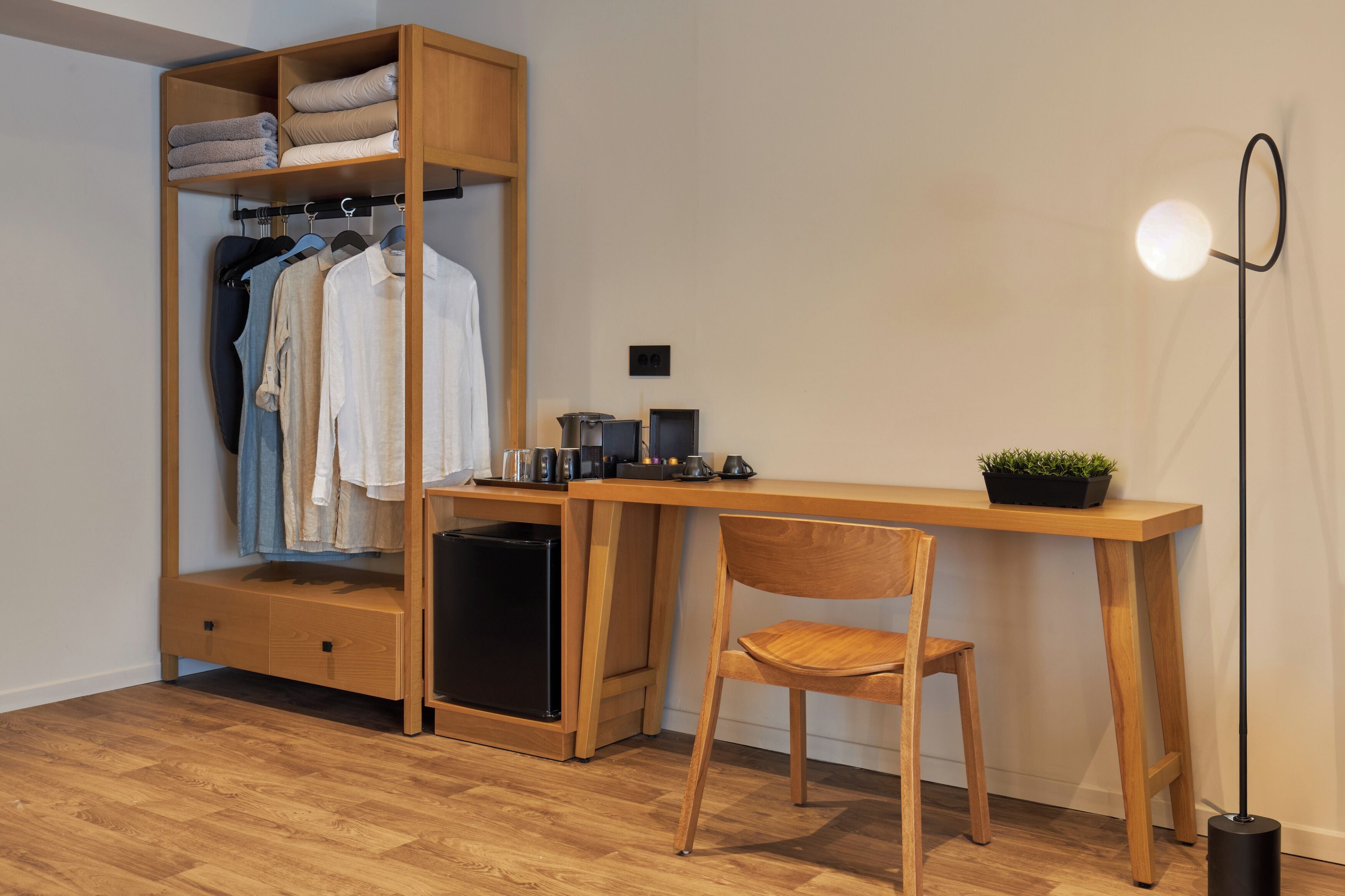A room with a wooden desk, a chair, a wardrobe with hanging clothes, a small black trash bin, and a floor lamp.