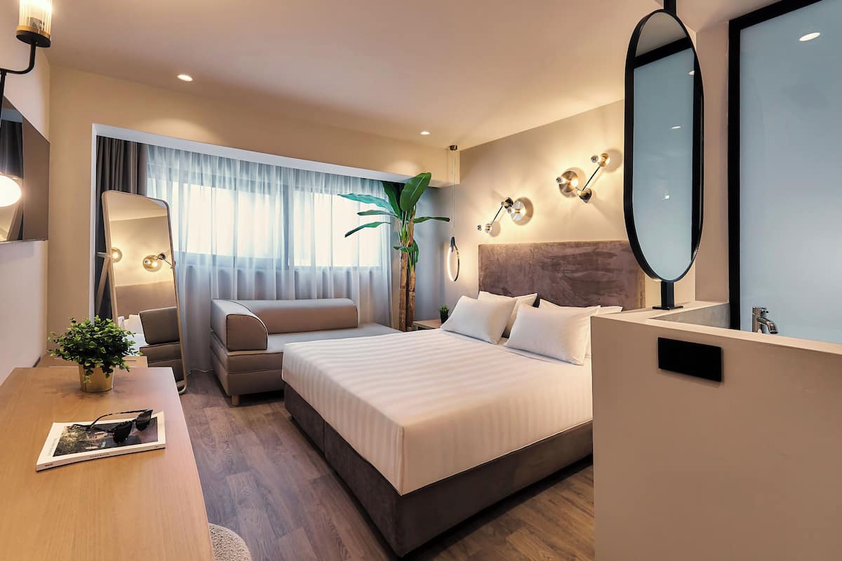 Una habitación de hotel moderna con una cama grande, un escritorio con una planta, un espejo y vistas al exterior.
