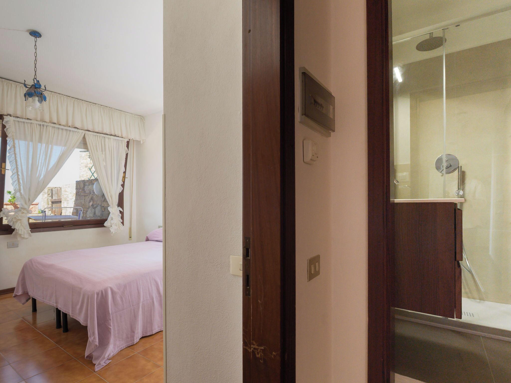 Ein Schlafzimmer mit Bett, einem Fenster mit Vorhängen, einer Duschkabine und einer Tür zu einem anderen Raum.
