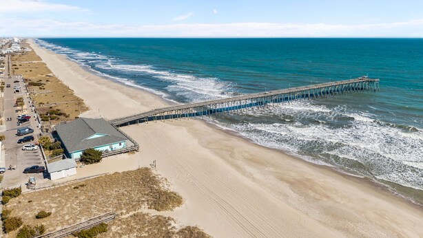 Ein Strand mit einer langen Pier, die sich ins Meer erstreckt, einem Parkplatz und einem Gebäude mit blauem Dach.