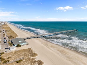 A beach with a long pier extending into the ocean, a parking area, and a building with a blue roof. - Kure Beach Villa - Walk to Beach, Pier & Dining, 2BR Condo w/ Pool (Kure Beach)