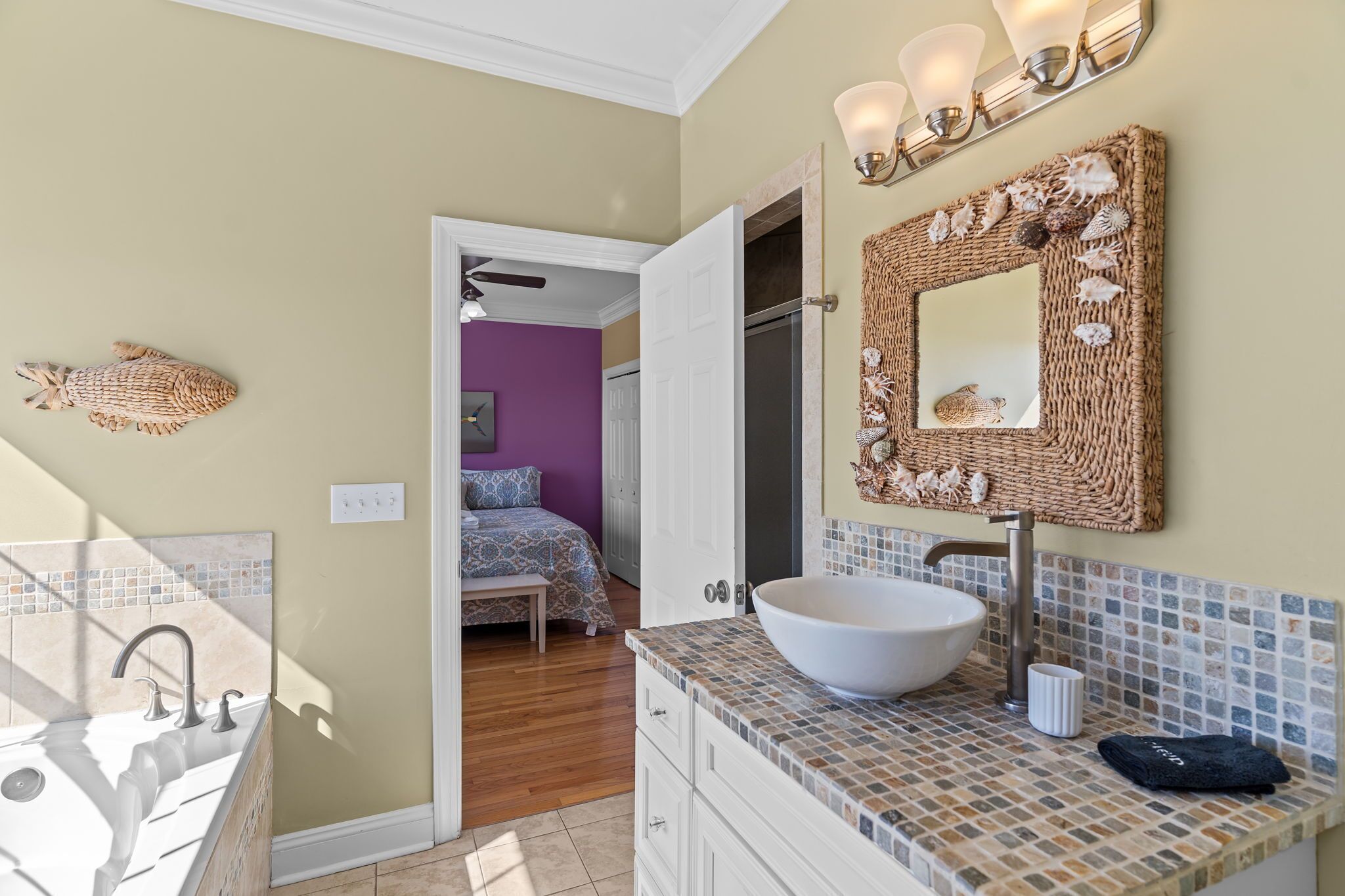 A bathroom with a large white vessel sink, a woven mirror frame with seashells, and a tiled backsplash.