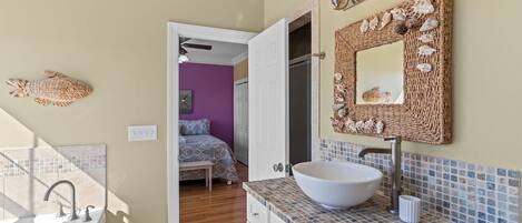 A bathroom with a large white vessel sink, a woven mirror frame with seashells, and a tiled backsplash.