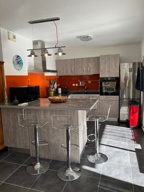 A modern kitchen with a central island, bar stools, stainless steel appliances, and a decorative clock on the wall. - 5 minutes from the beach, garden-level apartment with private parking. (La Ciotat)