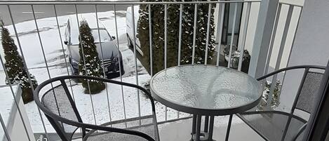 En balkong med ett metallbord och stolar, ett snötäckt område och utsikt över en byggnad med fönster.