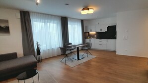 Ein modernes Wohnzimmer mit einer Couch, einem Esstisch mit Stühlen, einer Küchenzeile mit Schränken und einem großen Fenster mit Vorhängen.