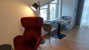 Ett modernt rum med en röd stol, ett litet bord, en lampa och utsikt över omgivningen genom stora fönster.