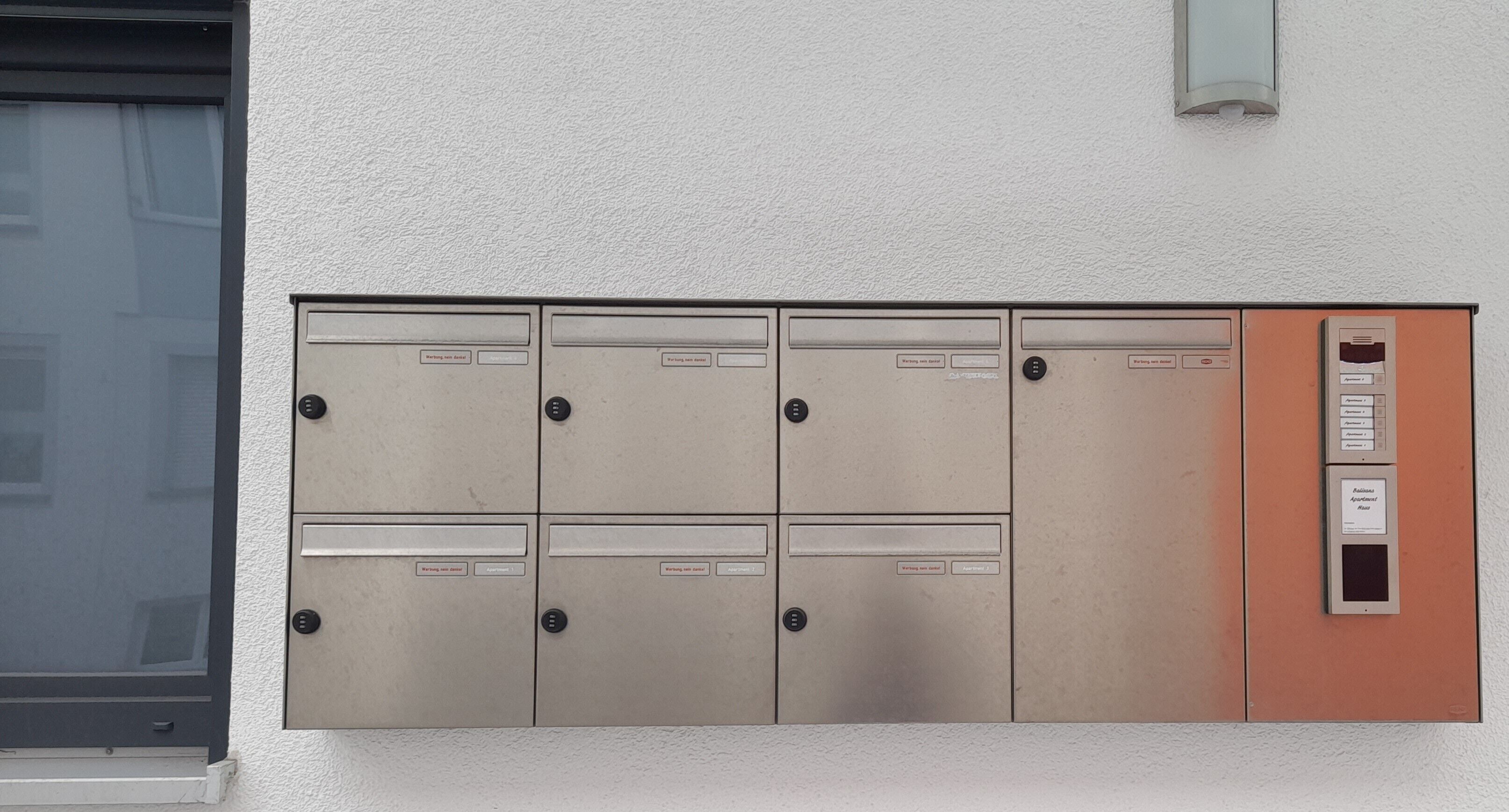 Un buzón empotrado en la pared con varios compartimentos, un pequeño panel naranja con botones y una lámpara encima.