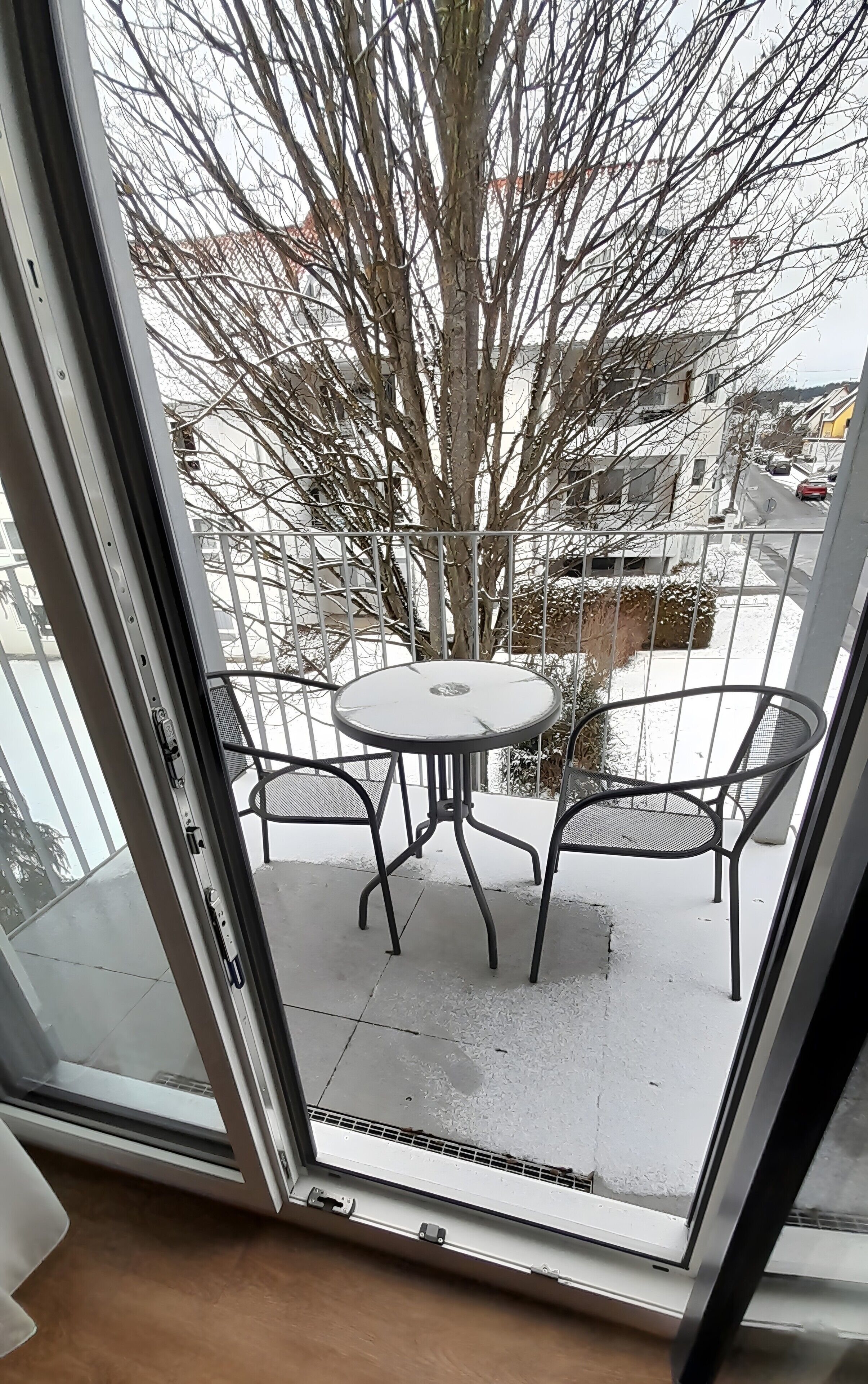 A view through a glass door to a balcony with a metal table and chairs, a tree, and a residential street.