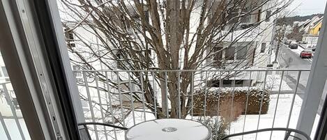 A view through a glass door to a balcony with a metal table and chairs, a tree, and a residential street.