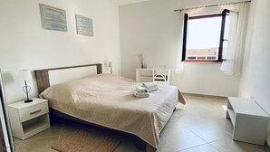 A bedroom with a bed, bedside tables, a nightstand, a window with curtains, and a view of buildings outside. - Vinko in Rovinj - Istrien / Vinko in Rovinj - Appartement für 2-3 Personen AP2 (Rovinj)