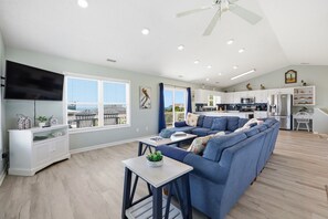 A modern living room with a sectional sofa, a flat-screen TV, and a dining area with a kitchen in the background. - Barefoot Pelican| Pool, Bikes & Game Room (Emerald Isle)