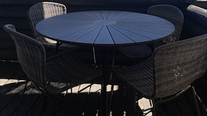 Mesa redonda con sillas de mimbre sobre una terraza de madera con vista a un lago, árboles y cielo despejado.