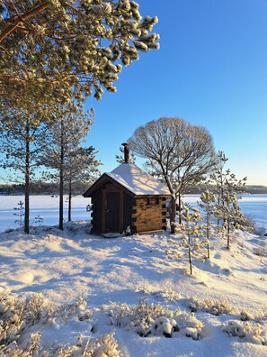 Eine Blockhütte mit schneebedecktem Dach, umgeben von schneebedeckten Bäumen und einem zugefrorenen See im Hintergrund.